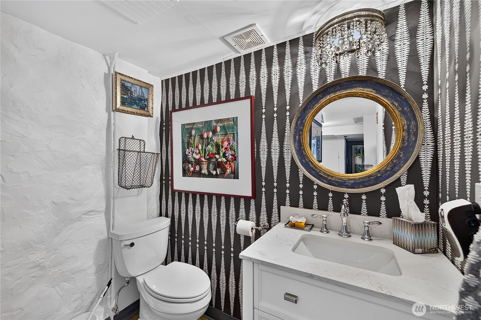 This powder room features a bold, high-contrast aesthetic with black and white patterned wallpaper and a textured white wall. The space is anchored by a vanity with a stone countertop, a circular mirror with a distressed blue and gold frame, and a crystal-style light fixture. A framed floral print and a wire wall basket add decorative character to this compact, stylish bathroom.