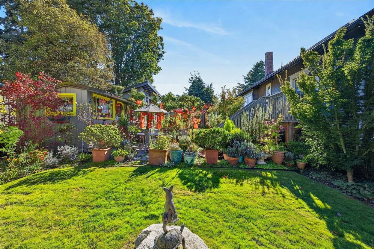 This vibrant backyard garden features a lush green lawn leading to a charming, eclectic outdoor living space filled with potted plants and hanging lanterns. A small, brightly accented shed sits to the left, while a wooden deck attached to the main house provides an elevated view of the colorful, well-manicured landscape. The scene is captured from a low, eye-level perspective, emphasizing the whimsical and inviting atmosphere of the private outdoor retreat.