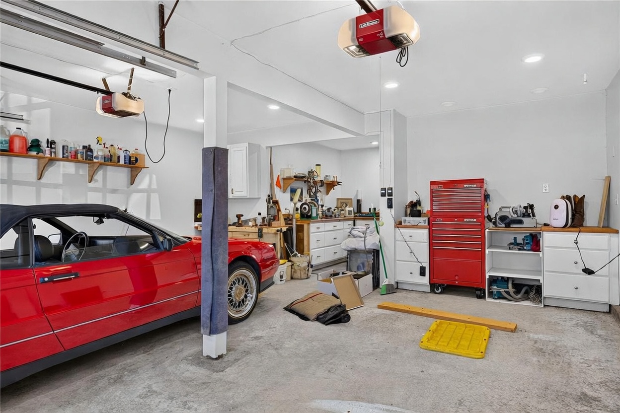 This spacious, well-lit garage features a red convertible parked on the left, with a dedicated workshop area in the back equipped with white cabinetry and a wooden workbench. The space includes a tall red rolling tool chest, ample shelving for storage, and two overhead garage door openers. The concrete floor and bright overhead lighting create a functional and organized environment for automotive maintenance or hobby projects.