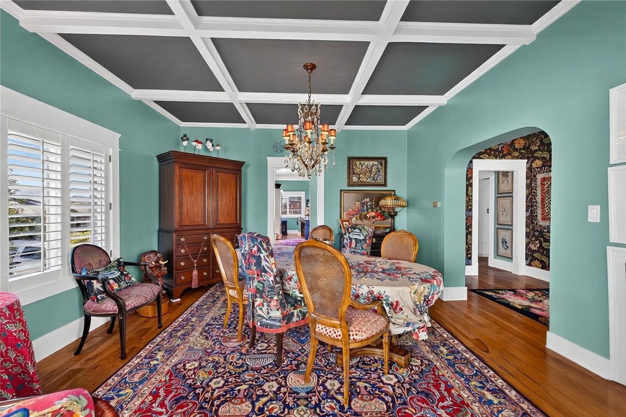 This formal dining room features a coffered ceiling with dark-painted panels and a decorative crystal chandelier. The space is furnished with a large oval table, patterned upholstered chairs, and a substantial wooden armoire, all set atop a vibrant, intricate area rug. An arched doorway leads to an adjacent room with floral wallpaper, creating a cohesive and traditional aesthetic.