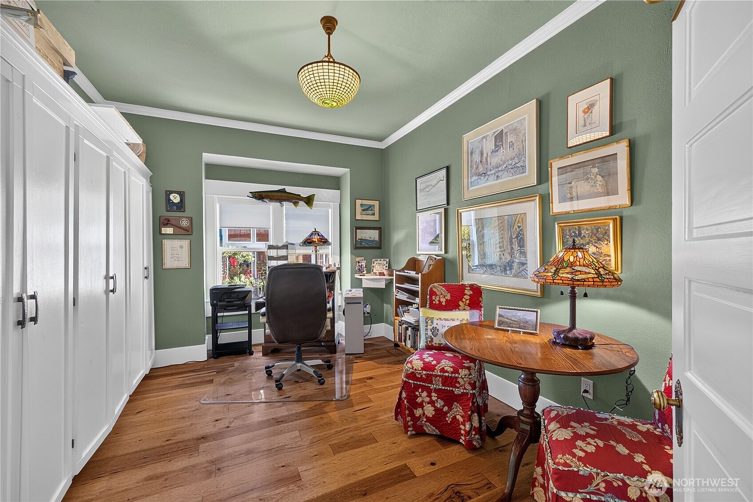 This cozy home office features sage green walls, warm wood flooring, and a large white storage unit along the left wall. A desk is positioned in front of a window, while a small round table with two patterned red chairs provides a comfortable seating area in the foreground. The room is decorated with numerous framed artworks and a stained-glass lamp, creating a traditional and personalized atmosphere.