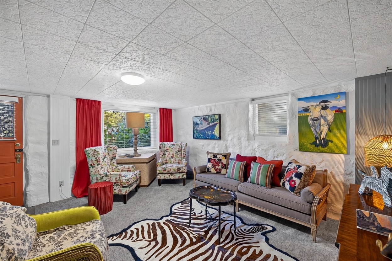 This eclectic living room features textured white stone walls and a drop ceiling, creating a cozy, basement-style atmosphere. The space is furnished with a grey sofa, two patterned wingback chairs, and a vibrant zebra-print area rug, complemented by colorful artwork and red curtains. A warm, inviting light is provided by a table lamp and a woven pendant fixture, making the room feel comfortable and lived-in.