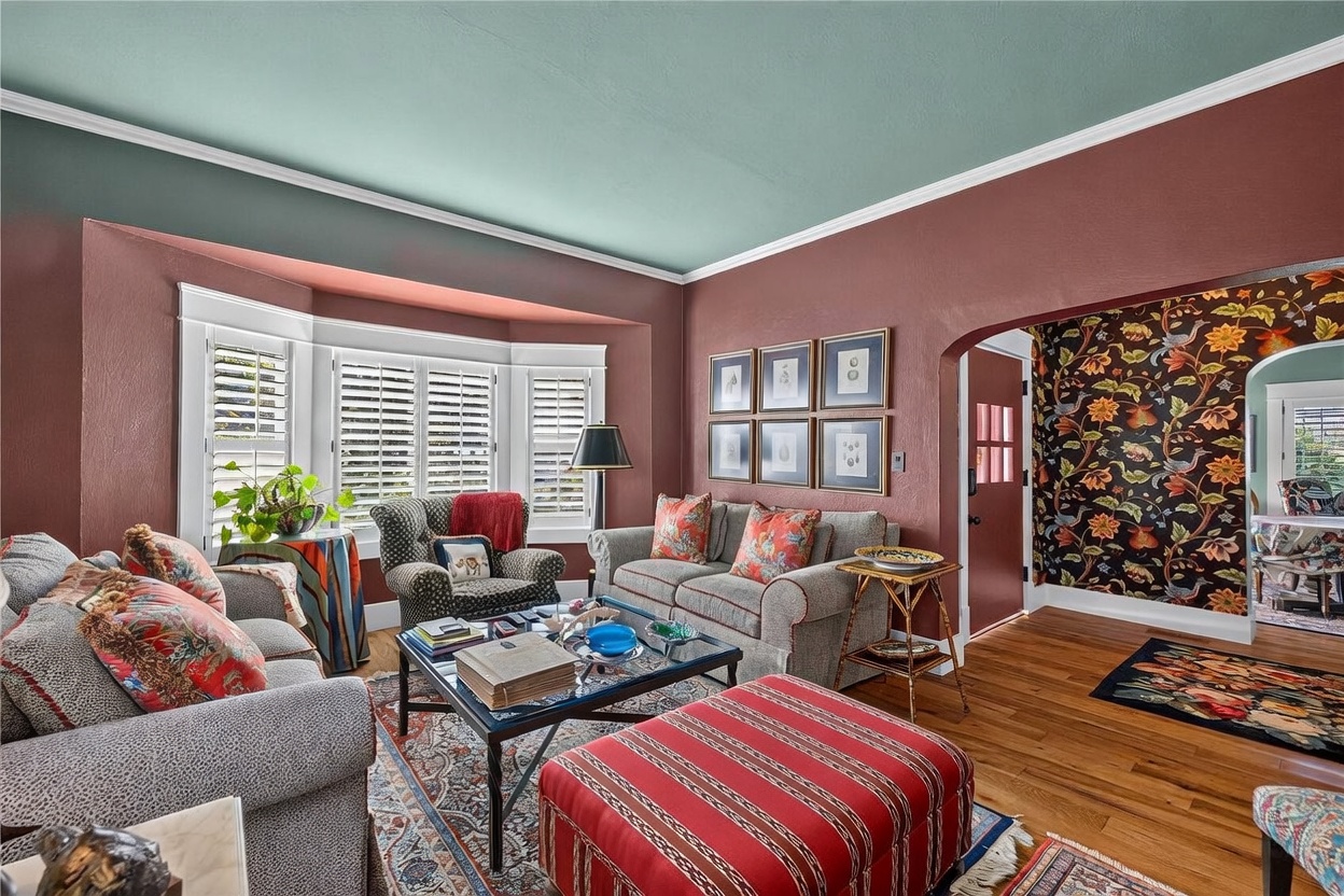 This cozy living room features deep, warm-toned walls contrasted by a soft teal ceiling and hardwood flooring. The space is furnished with patterned upholstered seating, a central coffee table, and a vibrant red striped ottoman, all anchored by a traditional area rug. A bay window with white shutters allows natural light to filter in, while an arched doorway leads to an adjacent room decorated with bold, floral wallpaper.