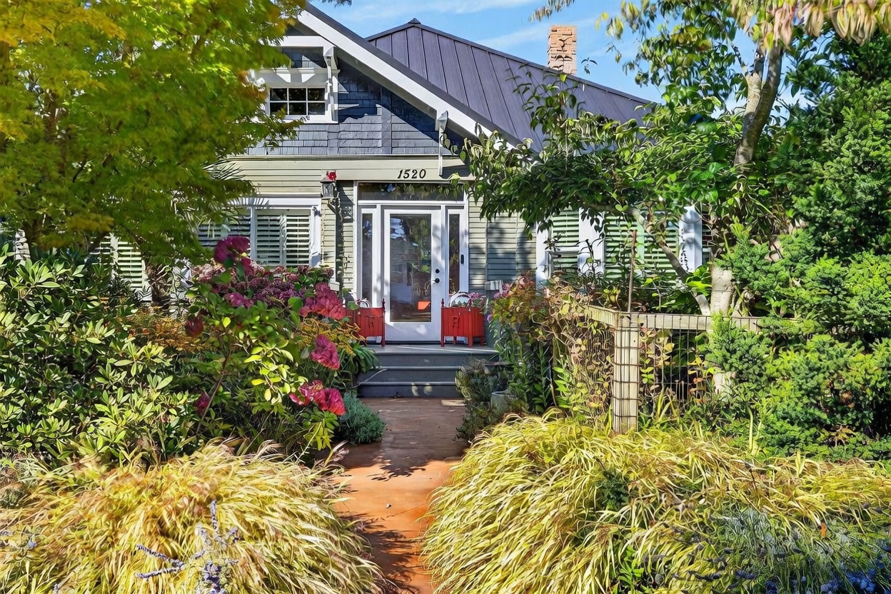 This charming cottage-style home features a classic front-facing gable with dark blue shingle siding and a metal roof. A lush, mature garden filled with vibrant hydrangeas and ornamental grasses leads up to a welcoming front entrance with a glass-paneled door. The perspective is a straight-on eye-level shot, capturing the inviting curb appeal and the well-maintained landscaping.