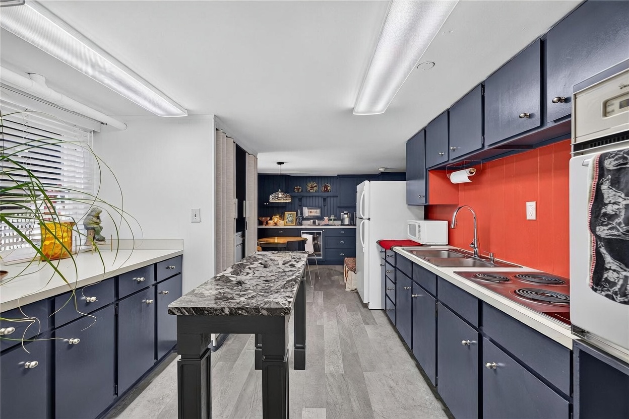 This kitchen features a bold design with dark blue cabinetry contrasted by a vibrant orange backsplash. A central island with a patterned stone countertop provides additional workspace, while the light-toned wood-look flooring adds a modern touch. The perspective looks through the galley-style kitchen toward a dining area in the background, creating a sense of depth and flow.