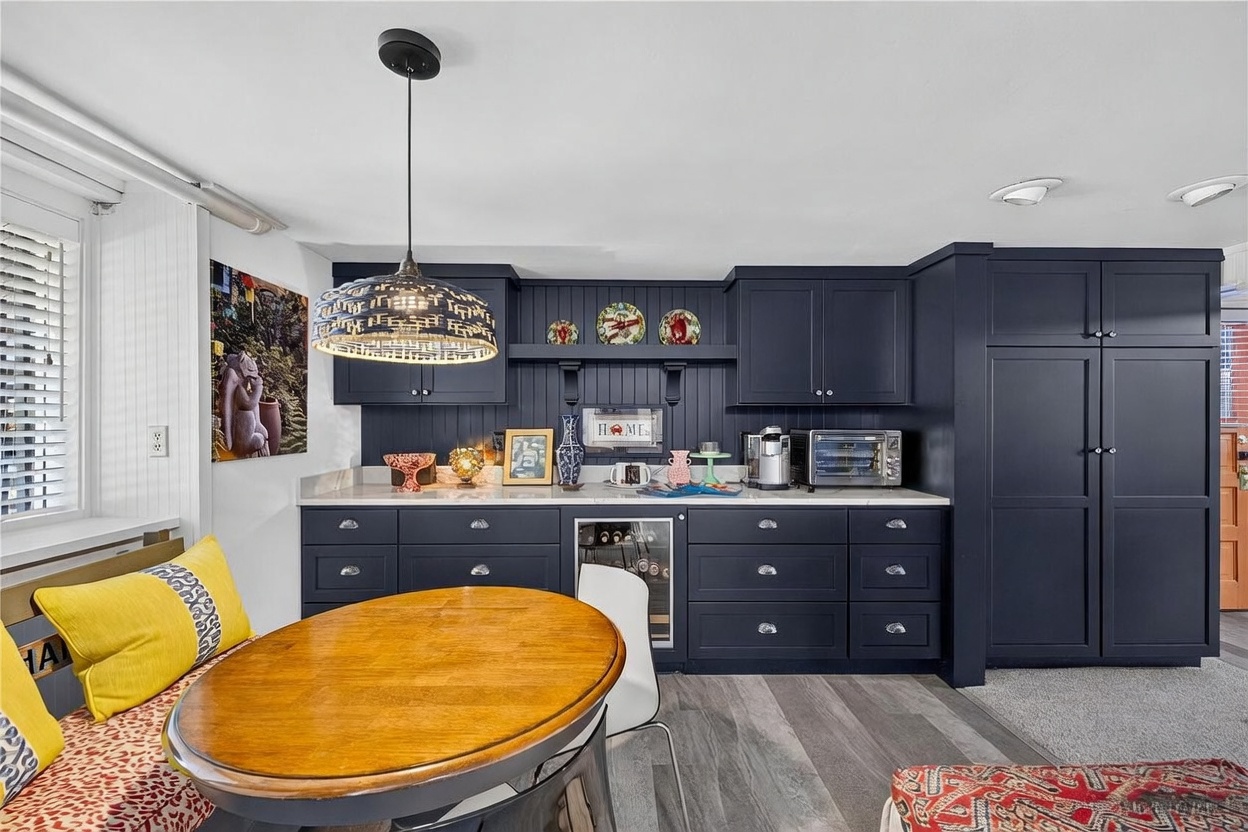 This kitchen area features dark navy cabinetry paired with a light-toned countertop and a matching backsplash. A central wooden dining table is surrounded by a built-in bench with patterned cushions, while a woven pendant light hangs above. The space includes a built-in wine cooler and a tall pantry cabinet, creating a functional and stylish breakfast nook or kitchenette.