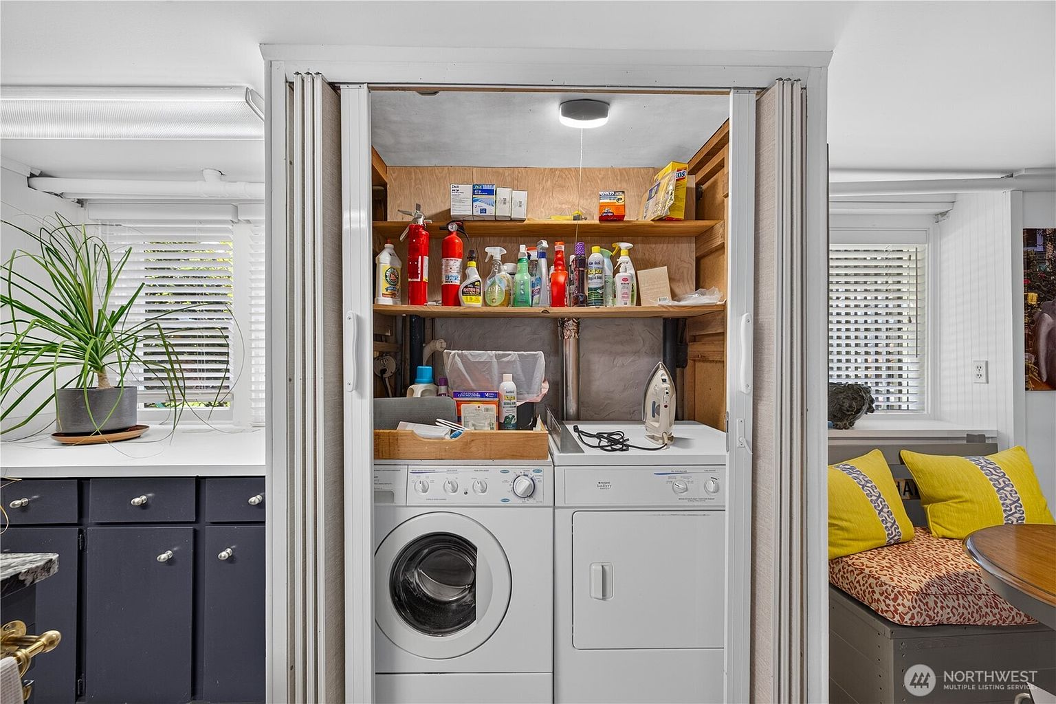 This laundry room features a stacked washer and dryer unit housed within a built-in closet space. Above the appliances, open shelving provides storage for cleaning supplies and household items, while a small iron rests on the dryer surface. The area is flanked by a dark-toned cabinet base on the left and a cozy seating nook with yellow cushions on the right, creating a functional and compact utility space.