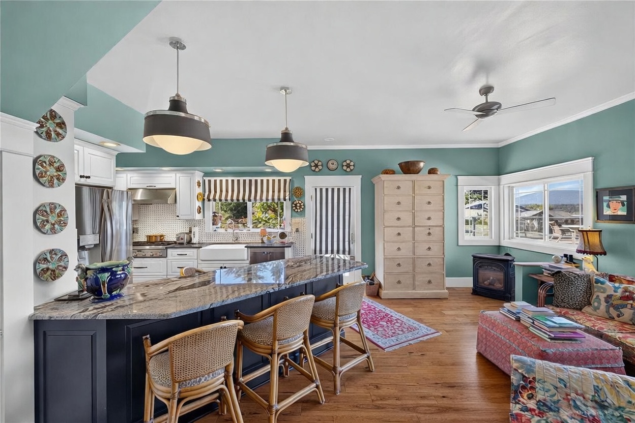 This open-concept kitchen features a large island with a dark base and a patterned stone countertop, complemented by three rattan bar stools. The space is accented by teal walls, white cabinetry, and a tall, light-colored wooden storage cabinet, creating a cozy and eclectic atmosphere. A small wood-burning stove sits in the corner near a window, while a patterned rug and decorative wall plates add character to the room.