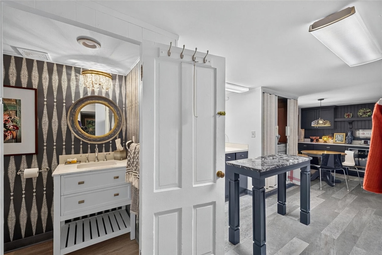 This image captures a view from a hallway looking into a powder room and an adjacent kitchenette area. The powder room features bold, patterned wallpaper, a circular mirror, and a vanity with a stone countertop, while the adjacent space includes a high-top table and a small kitchen setup with dark cabinetry. The perspective highlights the transition between the private bathroom space and the open-concept living area, showcasing a blend of decorative wallpaper and functional, modern finishes.