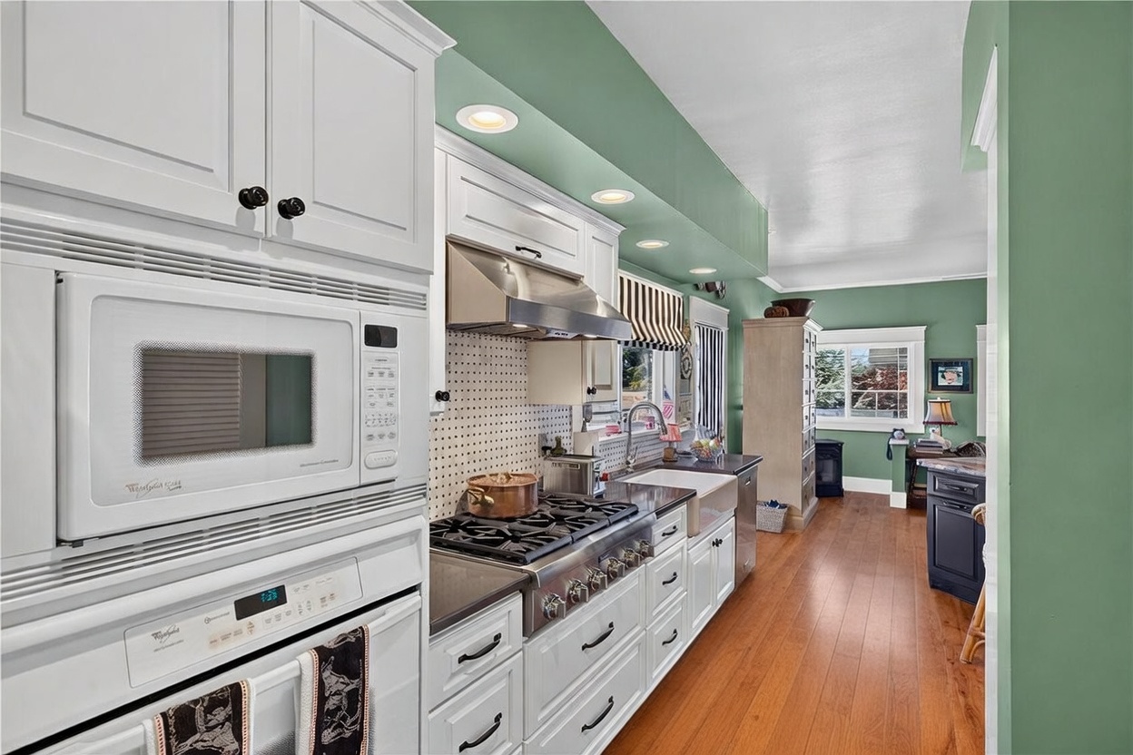This bright kitchen features white cabinetry paired with a stainless steel gas range and a built-in microwave oven. A farmhouse-style sink is set into dark countertops, complemented by a patterned tile backsplash and a soft green wall color. The perspective looks from the kitchen area toward an adjacent room, highlighting the hardwood flooring and a vibrant, patterned area rug that adds warmth to the space.