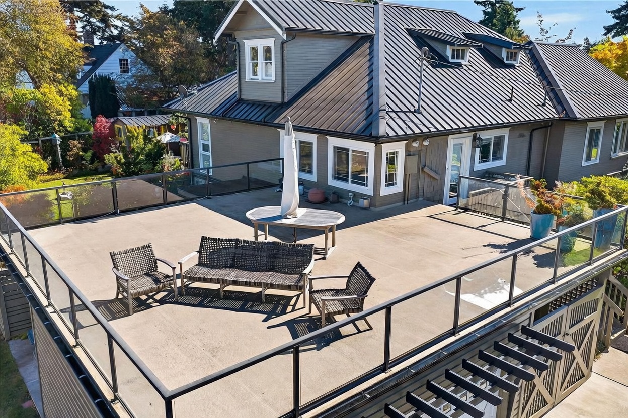 This elevated, expansive rooftop deck features a modern glass railing system and a neutral-toned surface, providing a clear view of the surrounding neighborhood. The space is furnished with a woven outdoor sofa set and a central dining table with a closed umbrella, creating an inviting area for relaxation or entertaining. The perspective is captured from an elevated angle, showcasing the home's dark metal roof, gray siding, and the seamless transition between the indoor living space and the outdoor terrace.