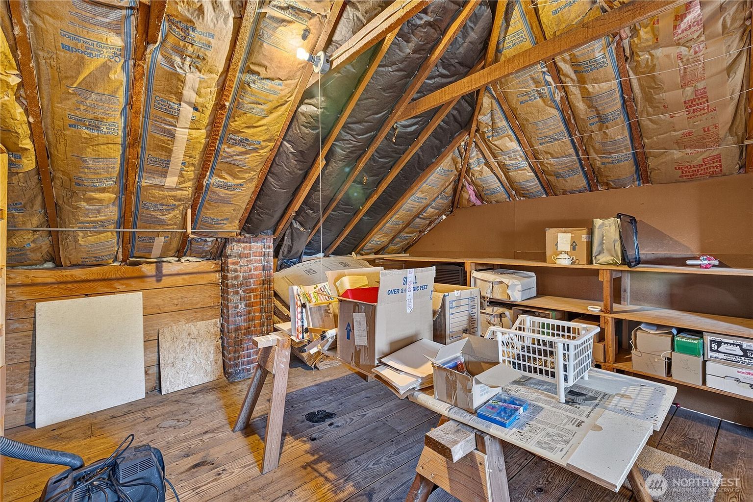This attic space features exposed wooden rafters and insulation batts tucked between the roof joists, creating a rustic and utilitarian atmosphere. The room is furnished with a wooden workbench, shelving units, and various storage boxes, suggesting a functional workshop or hobby area. A brick chimney stack rises through the space, adding a touch of character to the unfinished, wood-floored environment.