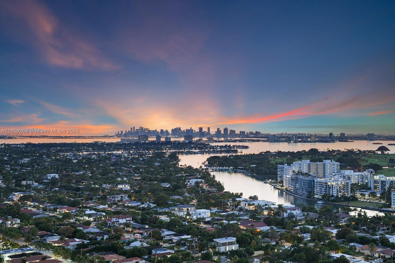 This aerial view showcases a sprawling residential area with numerous houses nestled among lush greenery, leading to a waterfront with a cityscape in the distance under a vibrant sunset sky. The scene captures a sense of community and tranquility, highlighting the proximity to both nature and urban amenities, making it an attractive location for potential homeowners.
