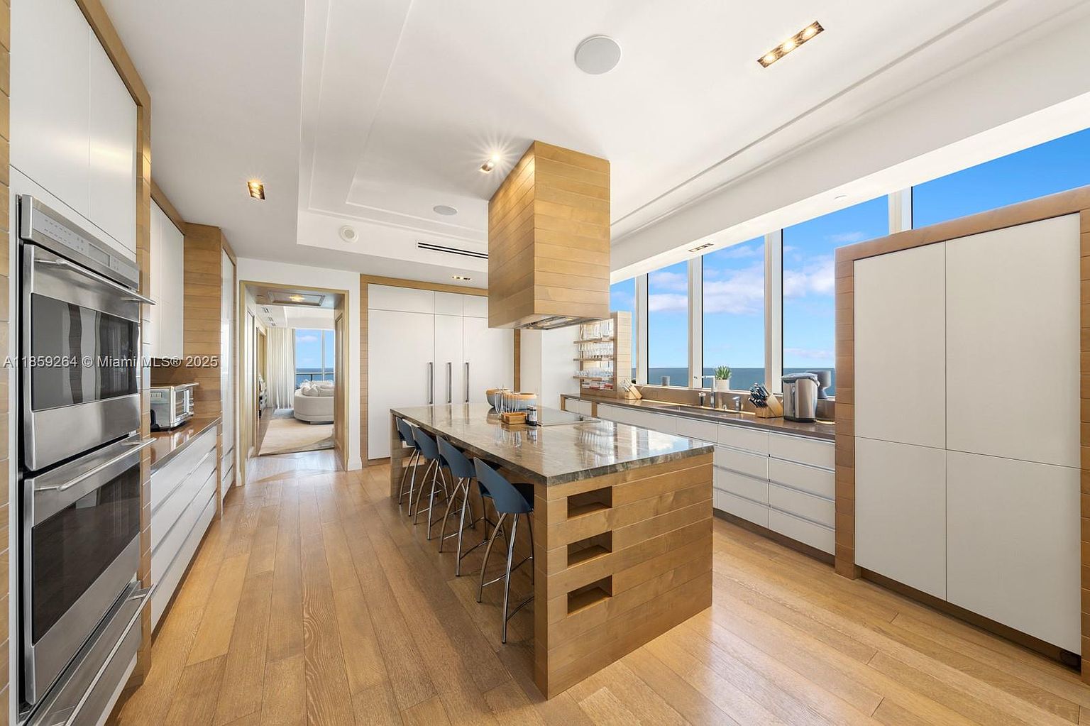 This is a bright and modern kitchen featuring a large wooden island with seating, stainless steel appliances, and white cabinetry with wooden trim. Large windows offer a view of the ocean, enhancing the luxurious feel of the space. The kitchen is spacious and well-lit, creating an inviting atmosphere.