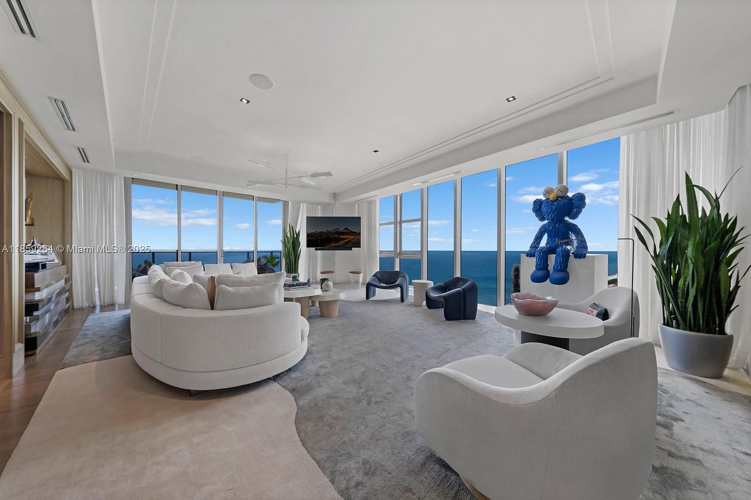 This is a spacious and luxurious living room with floor-to-ceiling windows offering stunning ocean views. The room features a large, curved white sofa, modern armchairs, and a unique blue sculpture, all arranged on a plush gray rug. The overall design is contemporary and elegant, creating a bright and inviting atmosphere.