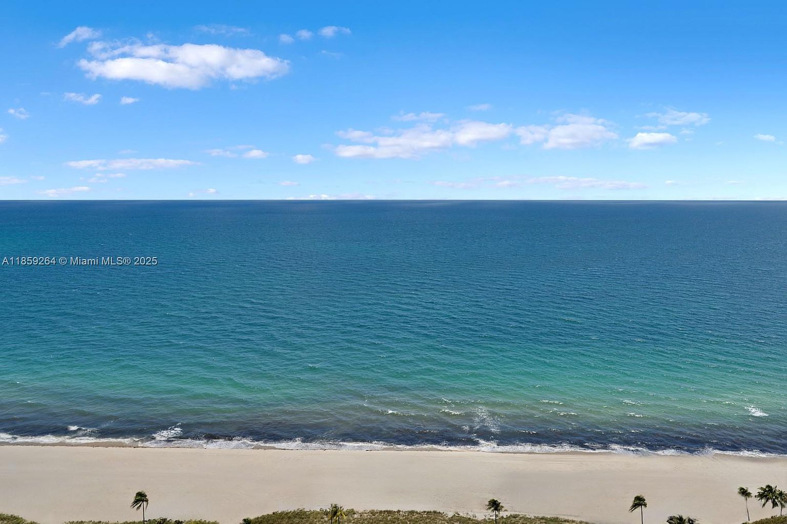 This aerial view showcases a stunning oceanfront property with a pristine sandy beach. The turquoise water transitions to a deep blue horizon under a clear sky dotted with fluffy clouds. Palm trees line the edge of the beach, adding a tropical touch to this idyllic coastal scene.