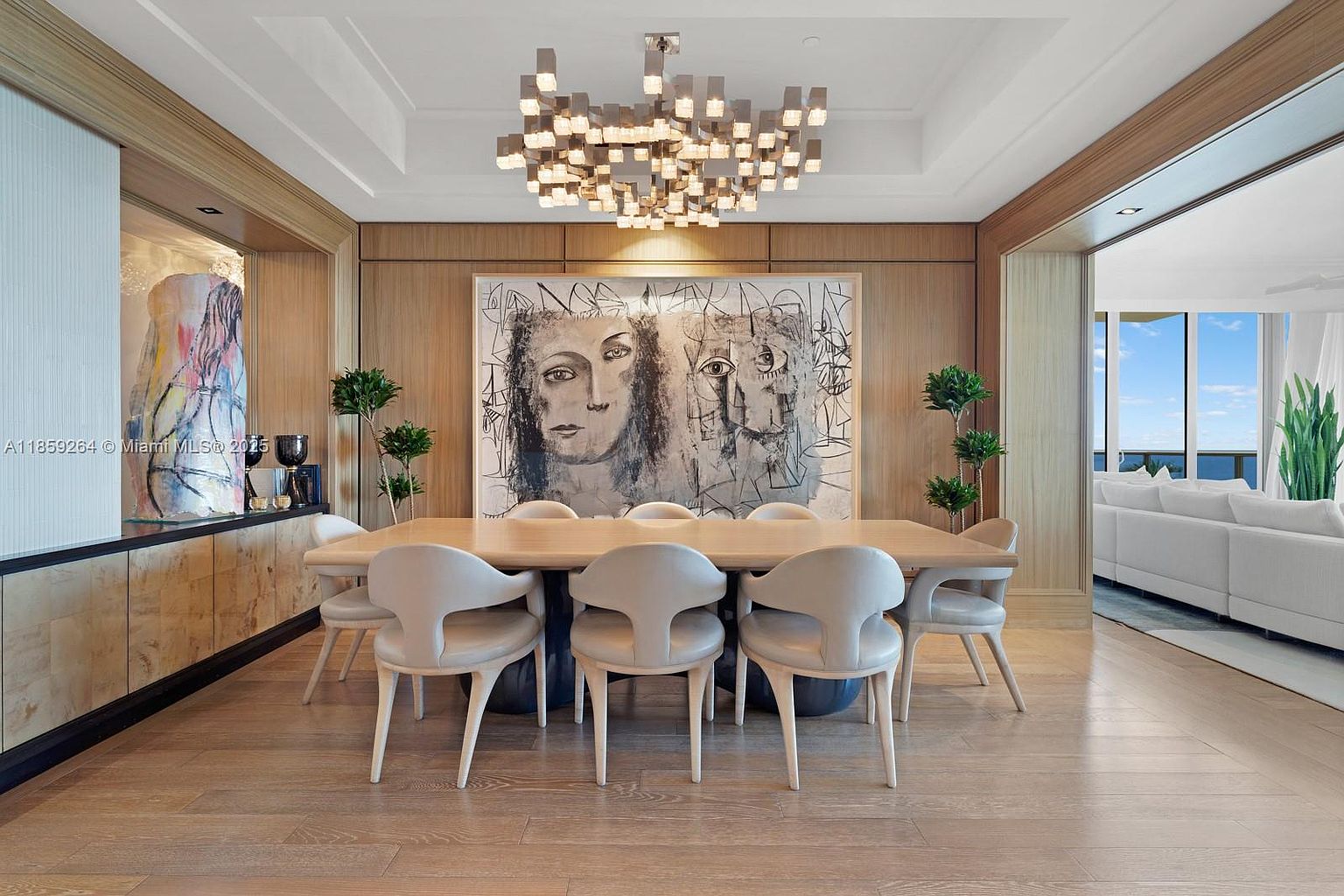 This is an interior shot of a dining room featuring a large wooden table surrounded by modern chairs with a unique design. Above the table hangs a contemporary chandelier, and a large abstract painting adorns the wall. The room has wood paneling and opens into a living area, creating a sense of spaciousness and luxury.