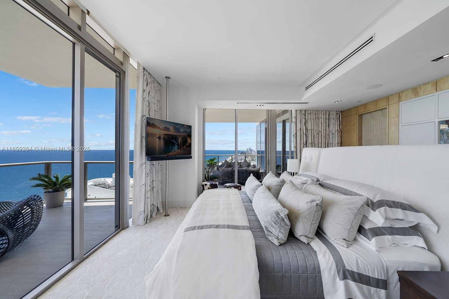 This is a luxurious primary bedroom with floor-to-ceiling windows offering stunning ocean views. The room features a large bed with plush pillows and a gray and white color scheme, complemented by elegant curtains and a wall-mounted television. The overall impression is one of comfort, sophistication, and high-end living.
