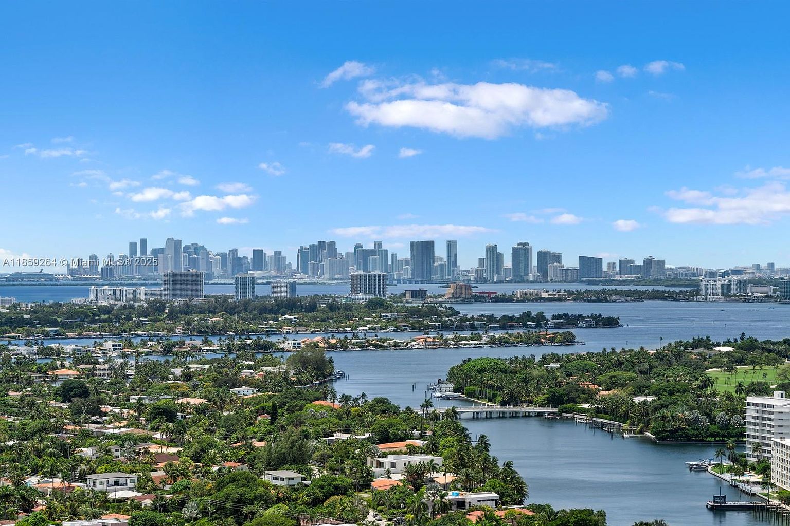 This aerial view showcases a stunning cityscape with a waterfront setting. Lush greenery and residential areas are visible in the foreground, leading to a bay with islands and bridges. In the distance, the skyline features numerous high-rise buildings, creating a vibrant urban backdrop under a partly cloudy sky.