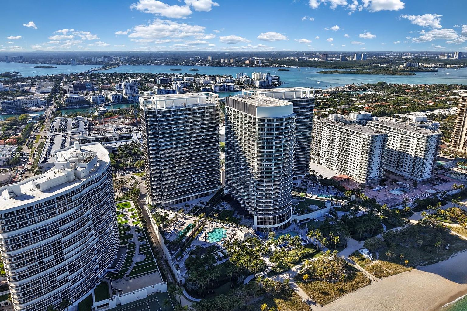 This aerial view showcases a luxury beachfront property featuring multiple high-rise condominium buildings with modern architectural designs. The buildings are surrounded by lush landscaping, including palm trees and manicured lawns, with a swimming pool area visible. The scene captures a vibrant coastal city with a beautiful waterfront and clear blue skies, emphasizing the upscale and desirable location.