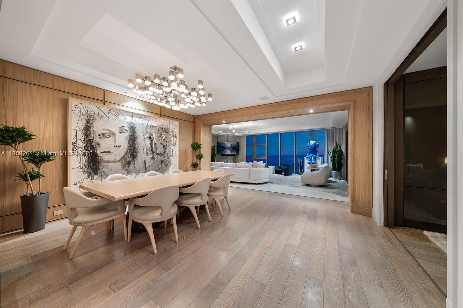 This interior shot showcases a luxurious dining room that seamlessly transitions into a living area with ocean views. The dining room features a large wooden table surrounded by modern chairs, illuminated by a statement chandelier, and complemented by a striking piece of art on the wall. The open layout and high-end finishes create an inviting and sophisticated atmosphere, perfect for entertaining.