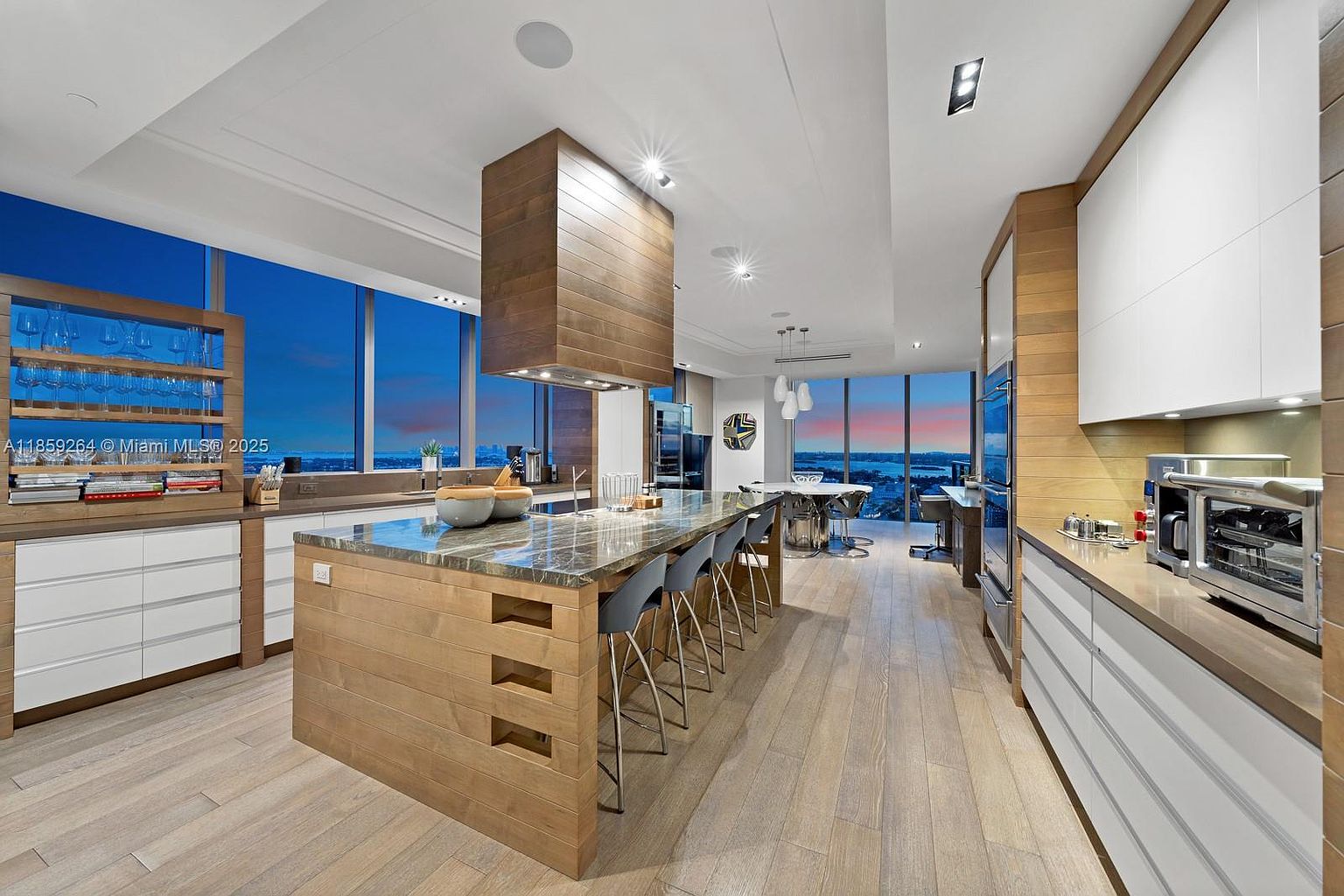 This is a modern, high-end kitchen featuring a large island with a marble countertop and seating, custom wood cabinetry, and stainless steel appliances. The kitchen has an open layout with large windows offering expansive views, and the hardwood flooring adds warmth to the space. The overall impression is luxurious and sophisticated.