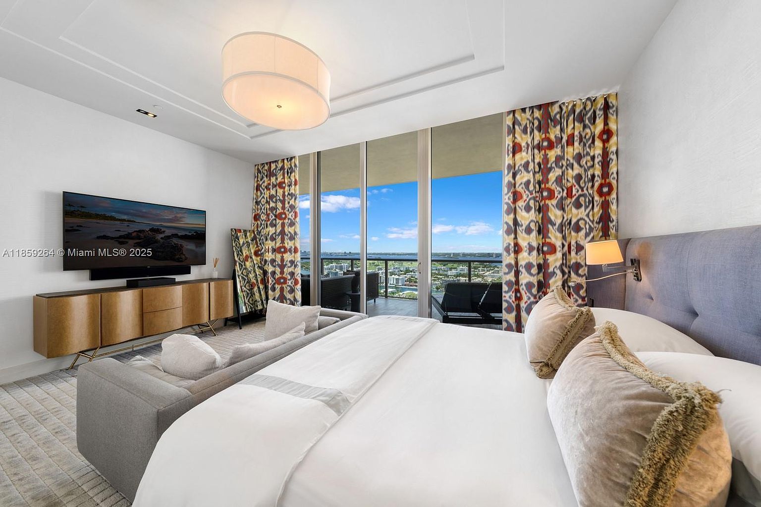 This is a primary bedroom featuring a king-size bed with white linens and decorative pillows. The room has floor-to-ceiling windows with patterned curtains, offering a view of the city skyline and water. A modern media console with a large television is positioned opposite the bed, and a gray sofa provides additional seating. The overall style is luxurious and contemporary.