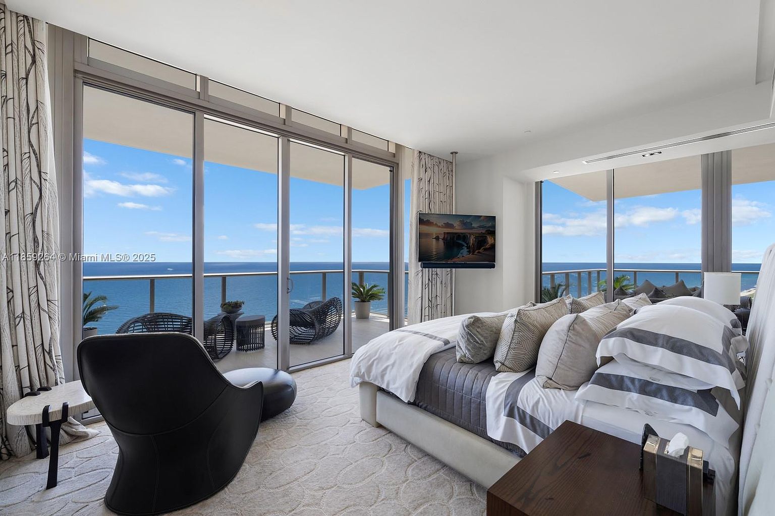 This is a luxurious primary bedroom featuring floor-to-ceiling windows with ocean views, a large bed with high-end linens, and a modern aesthetic. The room includes a sleek TV mounted on the wall and a stylish lounge chair, creating a relaxing and upscale atmosphere. The neutral color palette and natural light enhance the sense of tranquility and sophistication.