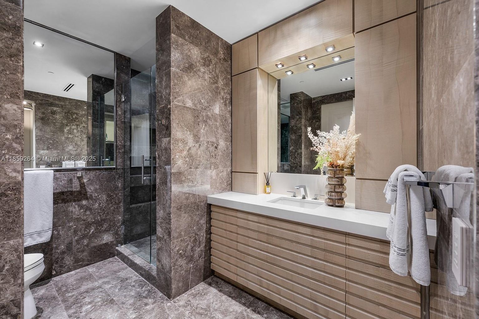 This is a luxurious primary bathroom featuring marble walls and flooring, a glass-enclosed shower, and a modern vanity with a white countertop and light wood cabinetry. A large mirror with integrated lighting enhances the space, and decorative elements like a vase with flowers and neatly arranged towels add to the sophisticated ambiance. The perspective is from the doorway, showcasing the entire bathroom.