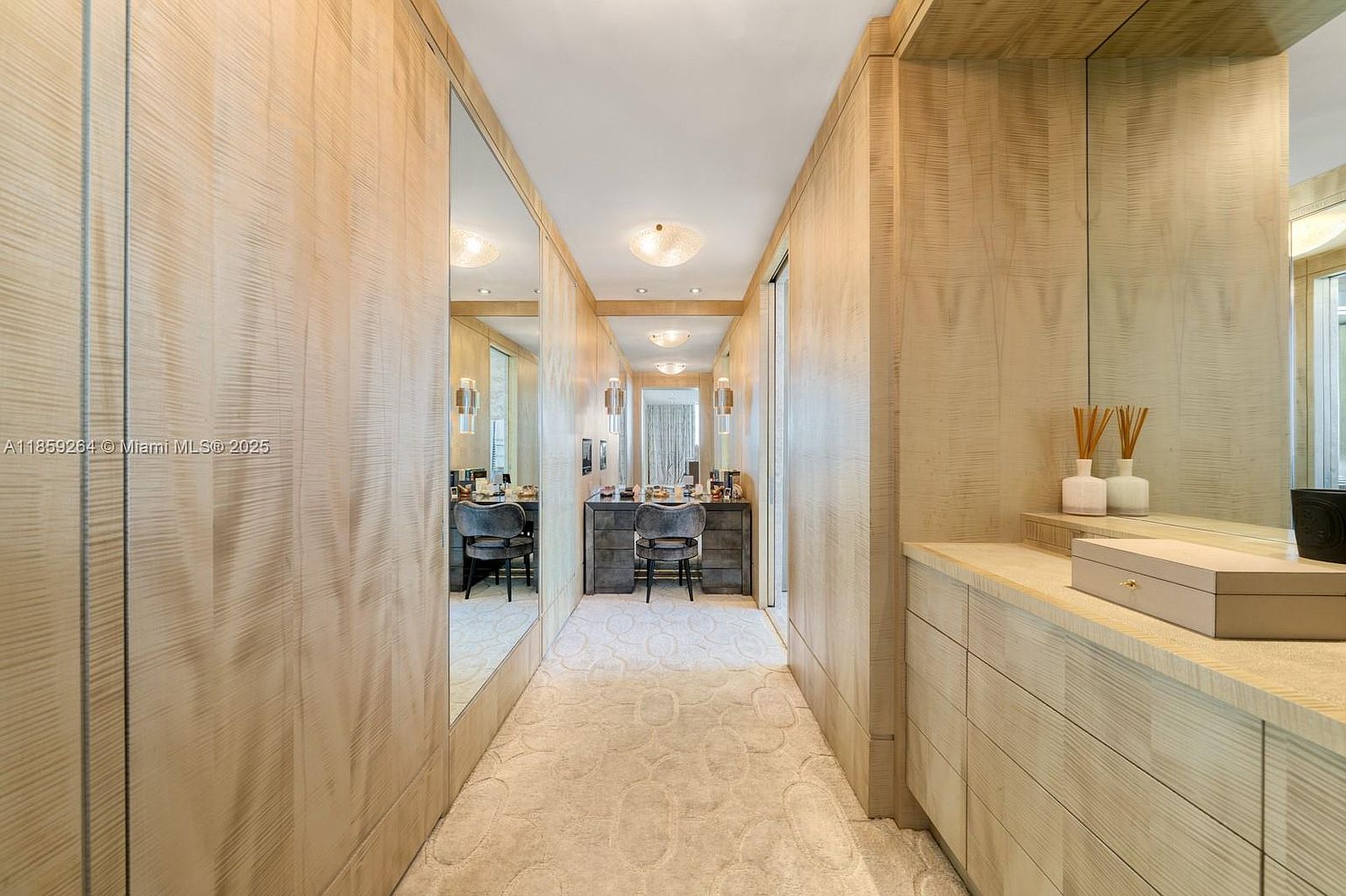 This is a well-organized walk-in closet featuring light wood paneling and mirrored walls that create an illusion of spaciousness. A vanity area with a chair is visible at the end of the closet, and the carpeted floor adds a touch of luxury. The closet is illuminated by ceiling lights and wall sconces.