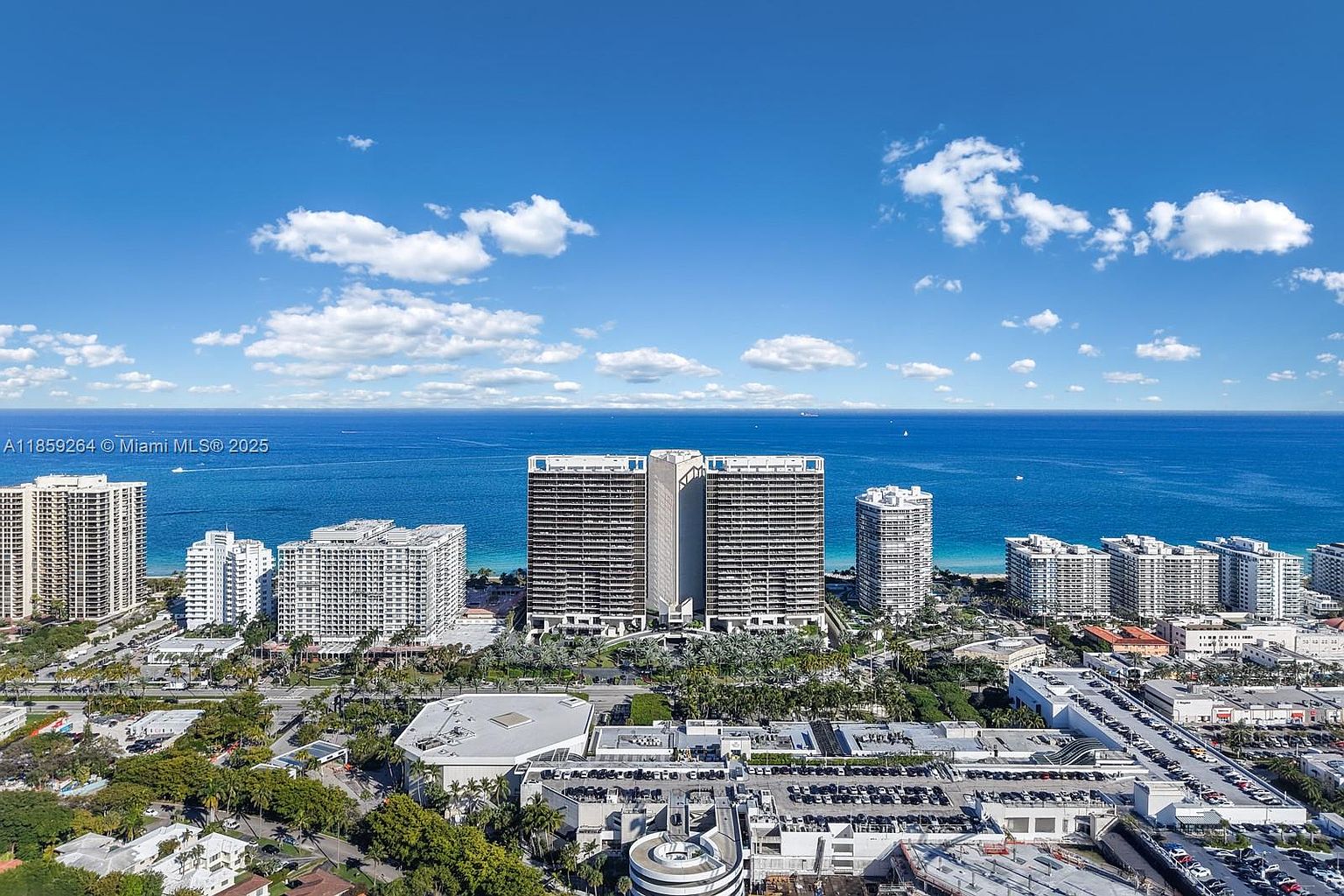 This aerial shot showcases a stunning coastal cityscape featuring multiple high-rise residential buildings against a backdrop of the ocean and a clear blue sky with scattered clouds. The buildings exhibit modern architectural styles, and the surrounding area includes lush greenery and well-maintained streets, creating an impression of luxury and urban living by the sea.