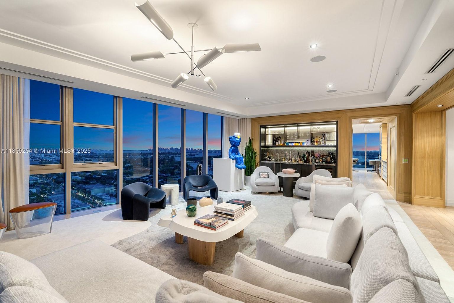This is a luxurious living room featuring floor-to-ceiling windows with a stunning city view. The room is furnished with a large, comfortable white sofa, stylish armchairs, and a unique coffee table. A built-in bar area adds a touch of sophistication, and the overall design is modern and elegant.