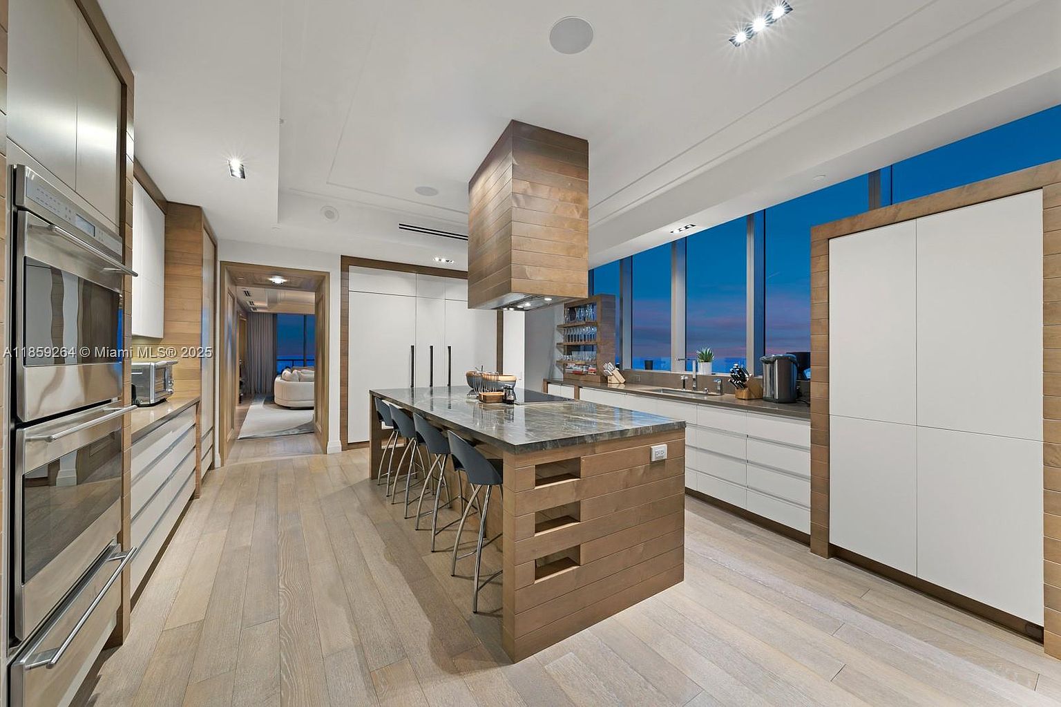 This is a modern, high-end kitchen featuring a large island with seating, sleek white cabinetry accented by wood trim, and stainless steel appliances. The kitchen is well-lit with recessed lighting and natural light from the large windows, which offer a view of the city skyline at dusk. The overall impression is one of luxury and sophistication.
