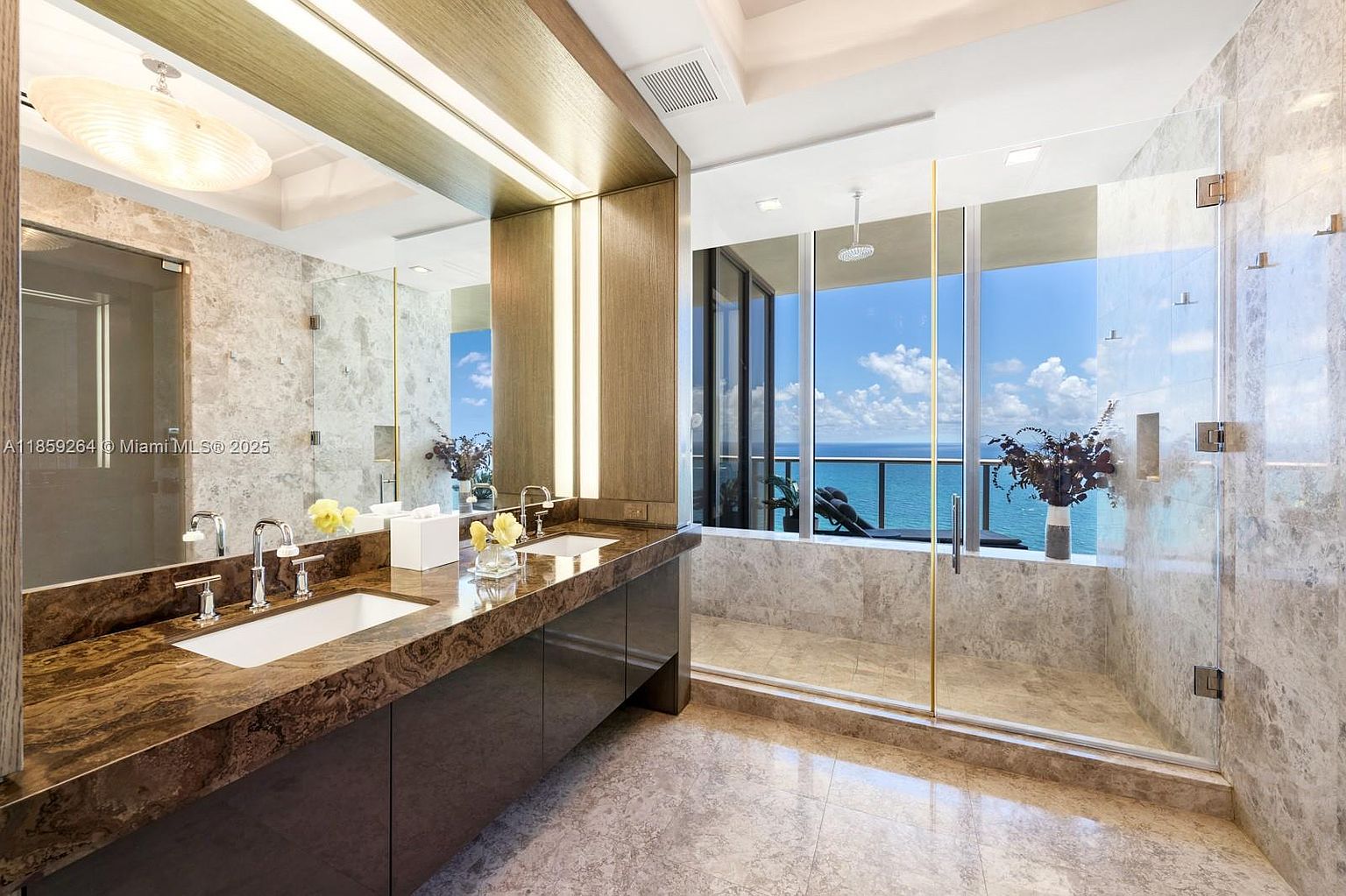 This is a luxurious primary bathroom featuring a double vanity with a brown marble countertop and modern fixtures. A large mirror with integrated lighting enhances the space. The bathroom includes a glass-enclosed shower with a stunning ocean view, creating a spa-like atmosphere. The floor and walls are covered in marble tiles, adding to the elegant design.