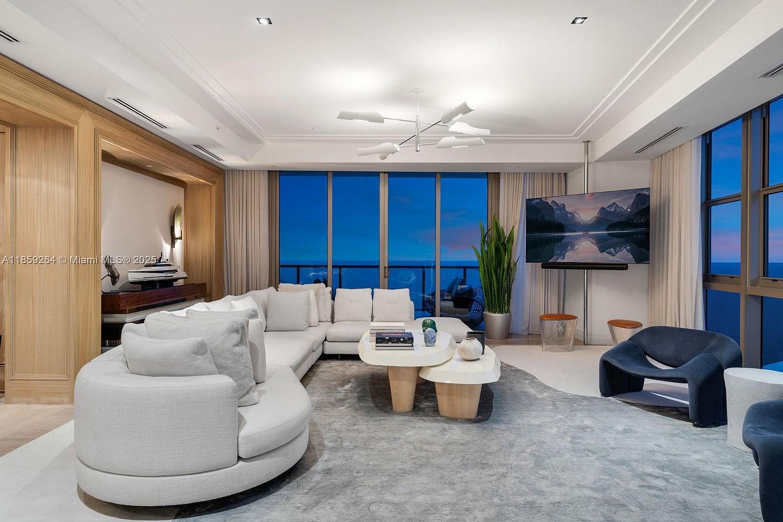 This is a luxurious living room featuring a large, curved white sofa, a modern chandelier, and floor-to-ceiling windows offering a stunning view. The room is decorated in neutral tones with a large area rug, creating a sophisticated and inviting atmosphere. A large television is mounted on a pole in front of the windows.