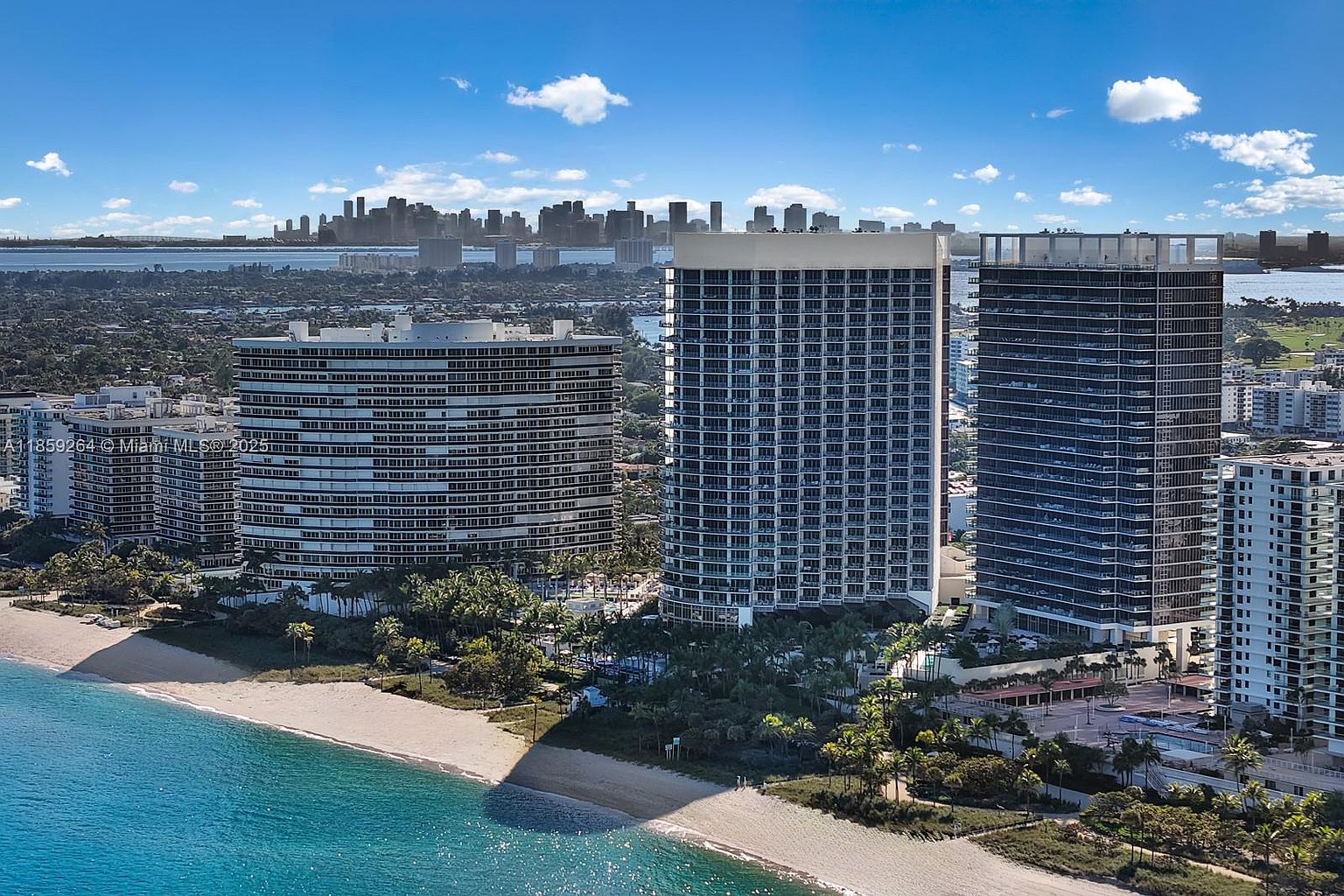 This aerial shot showcases a stunning beachfront property featuring multiple high-rise condominium buildings with a modern architectural style. The buildings are surrounded by lush greenery and pristine sandy beaches, with turquoise ocean waters in the foreground. In the distance, a city skyline adds to the allure of this luxurious coastal real estate.