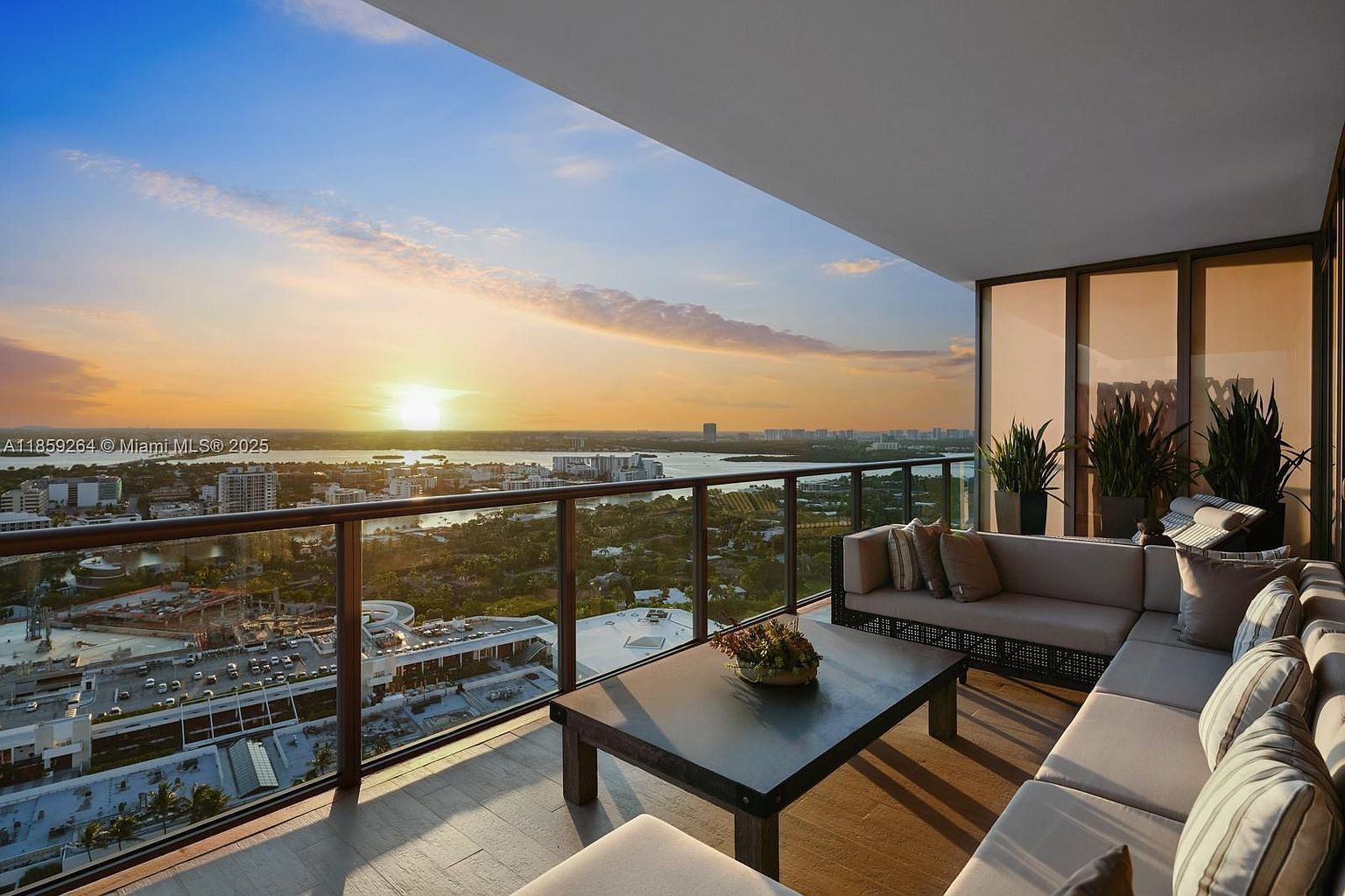 This image showcases a luxurious balcony with a stunning sunset view. The balcony features a comfortable sectional sofa with decorative pillows, a coffee table with a floral arrangement, and a glass railing that offers unobstructed views of the cityscape and waterfront. The overall impression is one of elegance and relaxation, perfect for enjoying the beautiful surroundings.