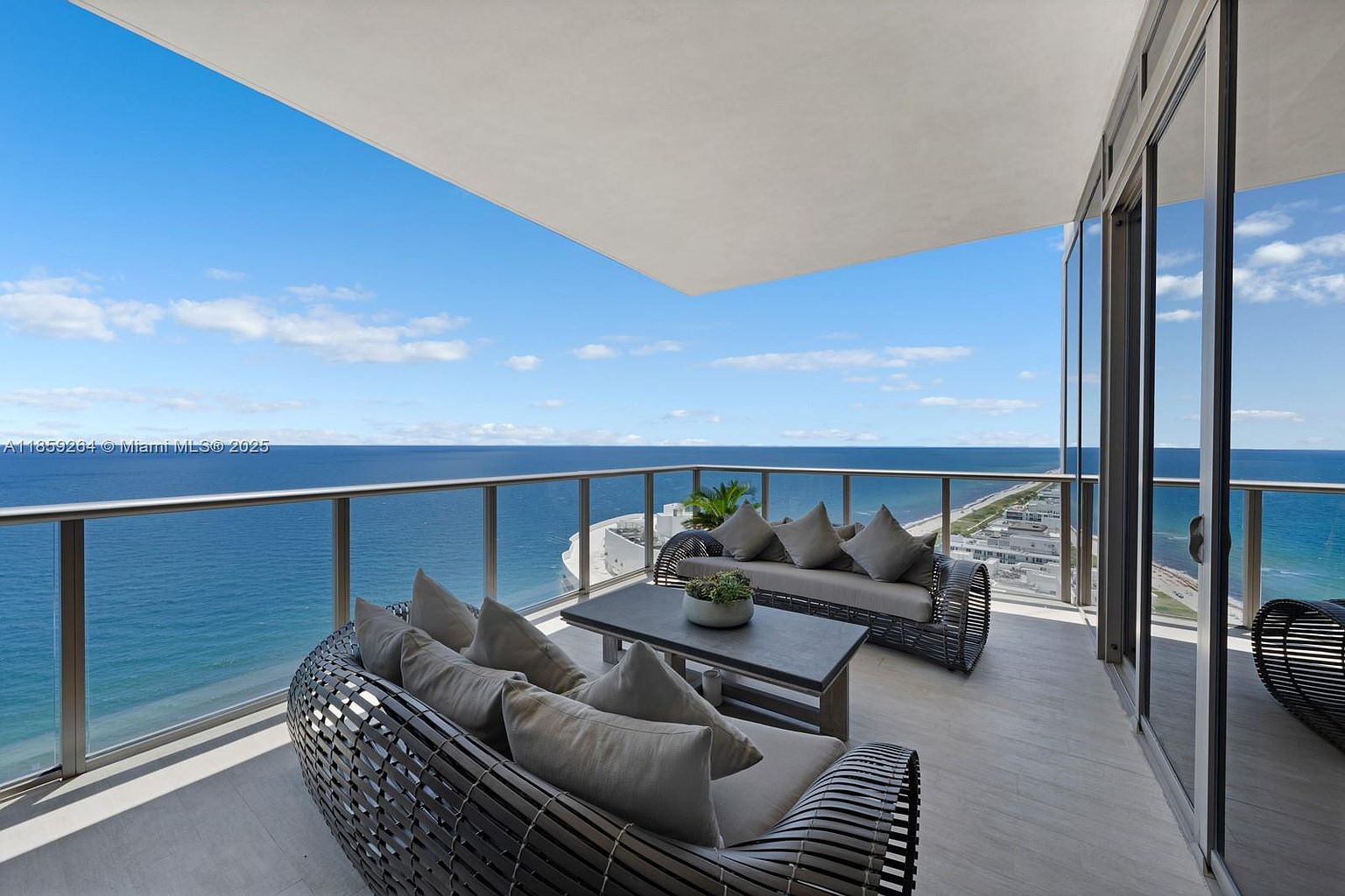 This image showcases a luxurious balcony with stylish outdoor furniture, including two woven sofas with plush cushions and a modern coffee table adorned with greenery. The balcony offers breathtaking panoramic views of the ocean and coastline, creating an inviting and serene outdoor living space. Large sliding glass doors seamlessly connect the indoor and outdoor areas, enhancing the sense of spaciousness and light.
