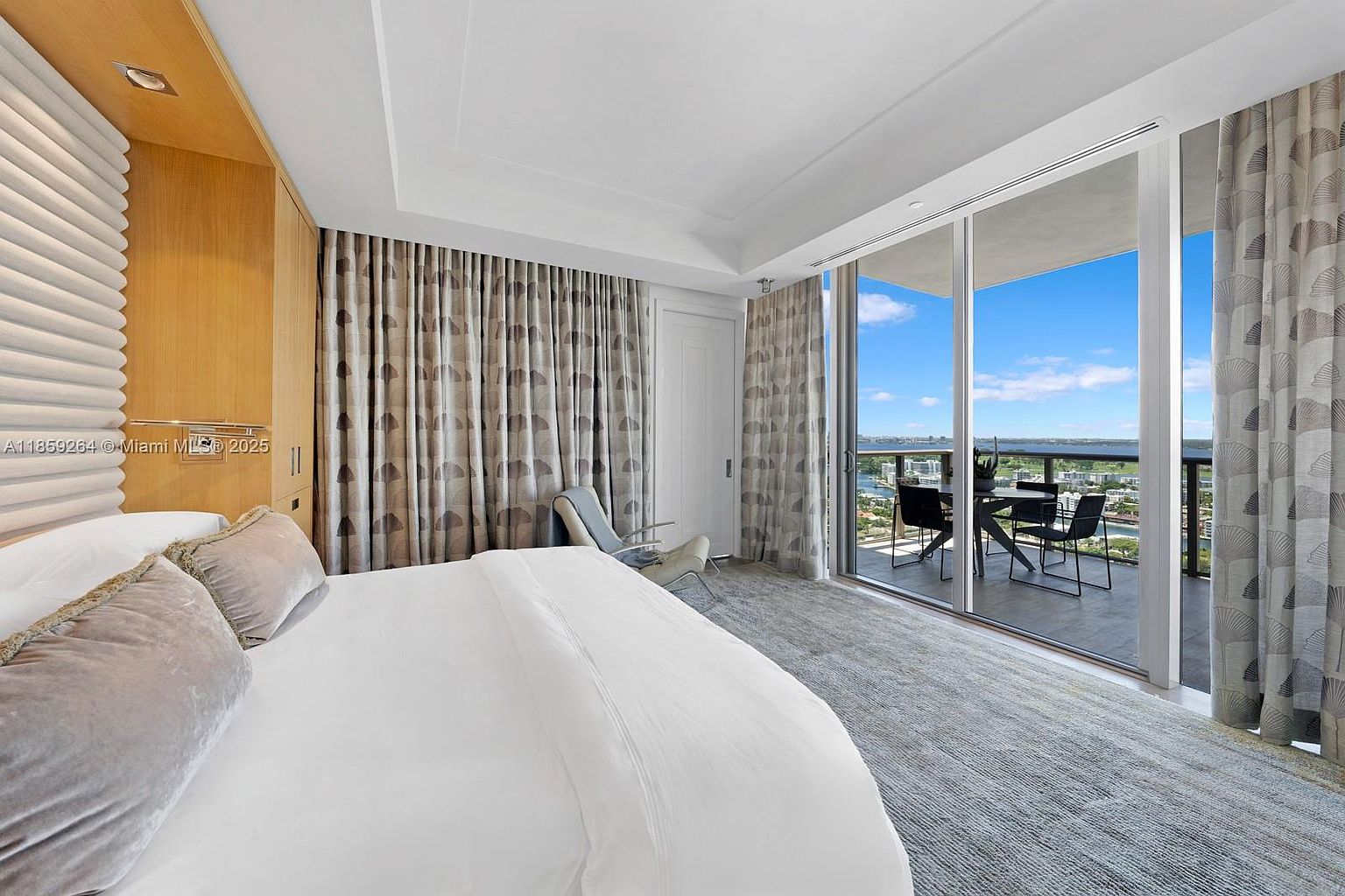 This is a primary bedroom featuring a large bed with plush pillows and a white comforter. The room has floor-to-ceiling curtains and a sliding glass door that leads to a balcony with outdoor seating and a view of the water and city. The overall style is modern and luxurious.