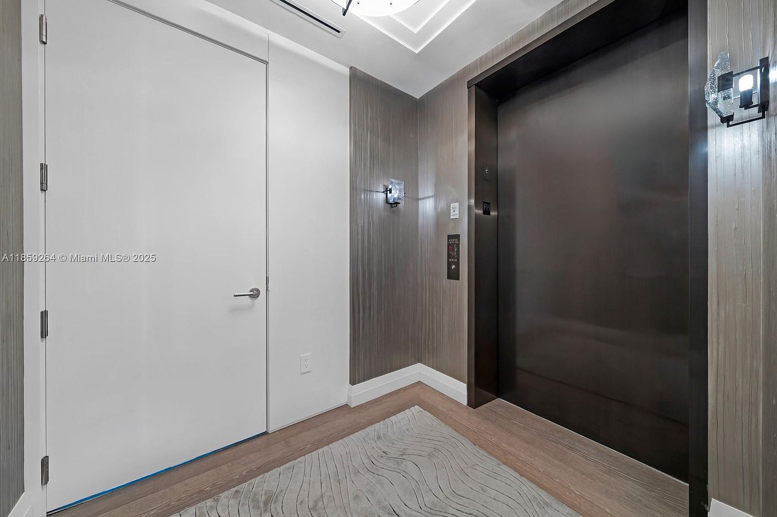 The image showcases a modern hallway featuring a sleek elevator and a contrasting white door. The walls are adorned with textured wallpaper, complemented by contemporary lighting fixtures. The flooring is light wood, adding warmth to the space, while a patterned rug adds a touch of elegance.