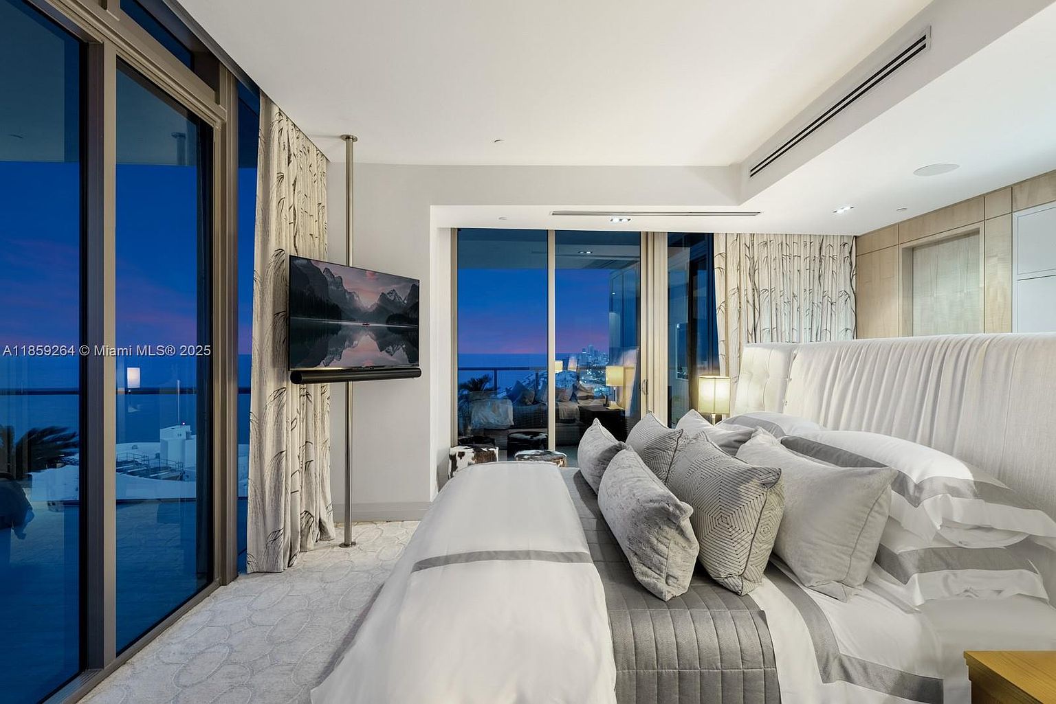 This is a luxurious primary bedroom featuring a large bed with plush pillows and a gray and white color scheme. A flat-screen TV is mounted on a stand, and floor-to-ceiling windows offer stunning views of the ocean and city skyline at dusk. The room exudes a modern and sophisticated ambiance.