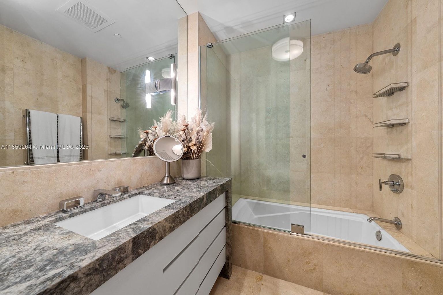 This is a well-lit primary bathroom featuring a granite countertop vanity with a white sink and modern faucet. A large mirror reflects the vanity and a portion of the adjacent shower/tub area, which is enclosed by glass. The walls and flooring are finished with light-colored stone tiles, creating a clean and luxurious aesthetic.