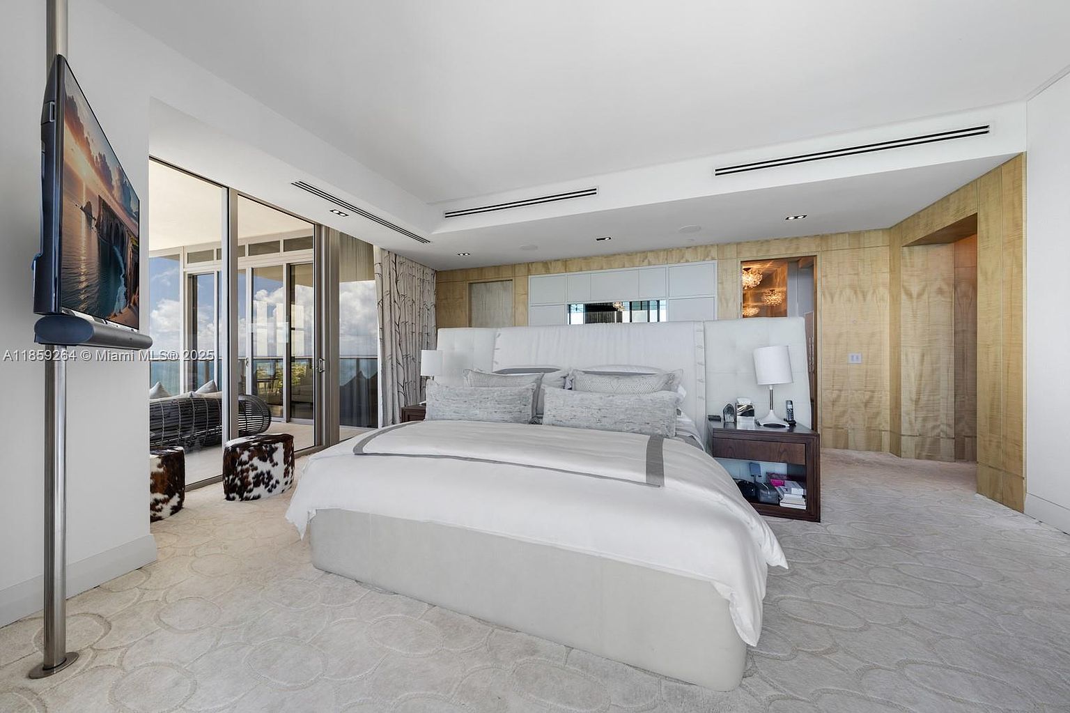 This is a luxurious primary bedroom featuring a large bed with a white headboard and plush bedding. The room has a neutral color palette with a patterned carpet and wood-paneled walls. A flat-screen TV is mounted on the wall, and there is access to a balcony with outdoor seating.