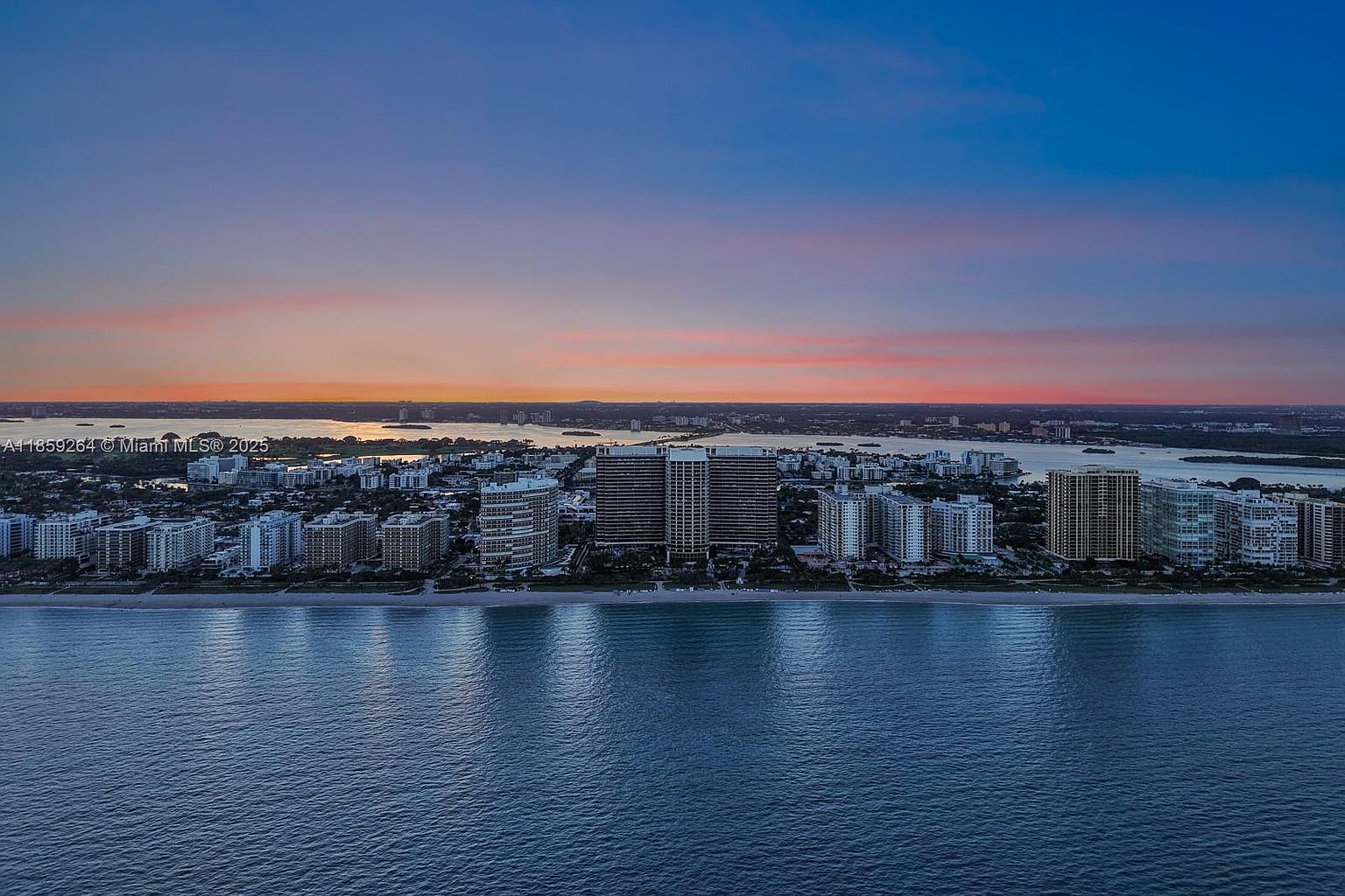 This aerial view showcases a stunning coastal cityscape at dusk. The scene features a row of high-rise buildings lining the beach, with the ocean in the foreground reflecting the colors of the sunset. The sky transitions from blue to orange and pink, creating a picturesque and desirable setting for luxury real estate.