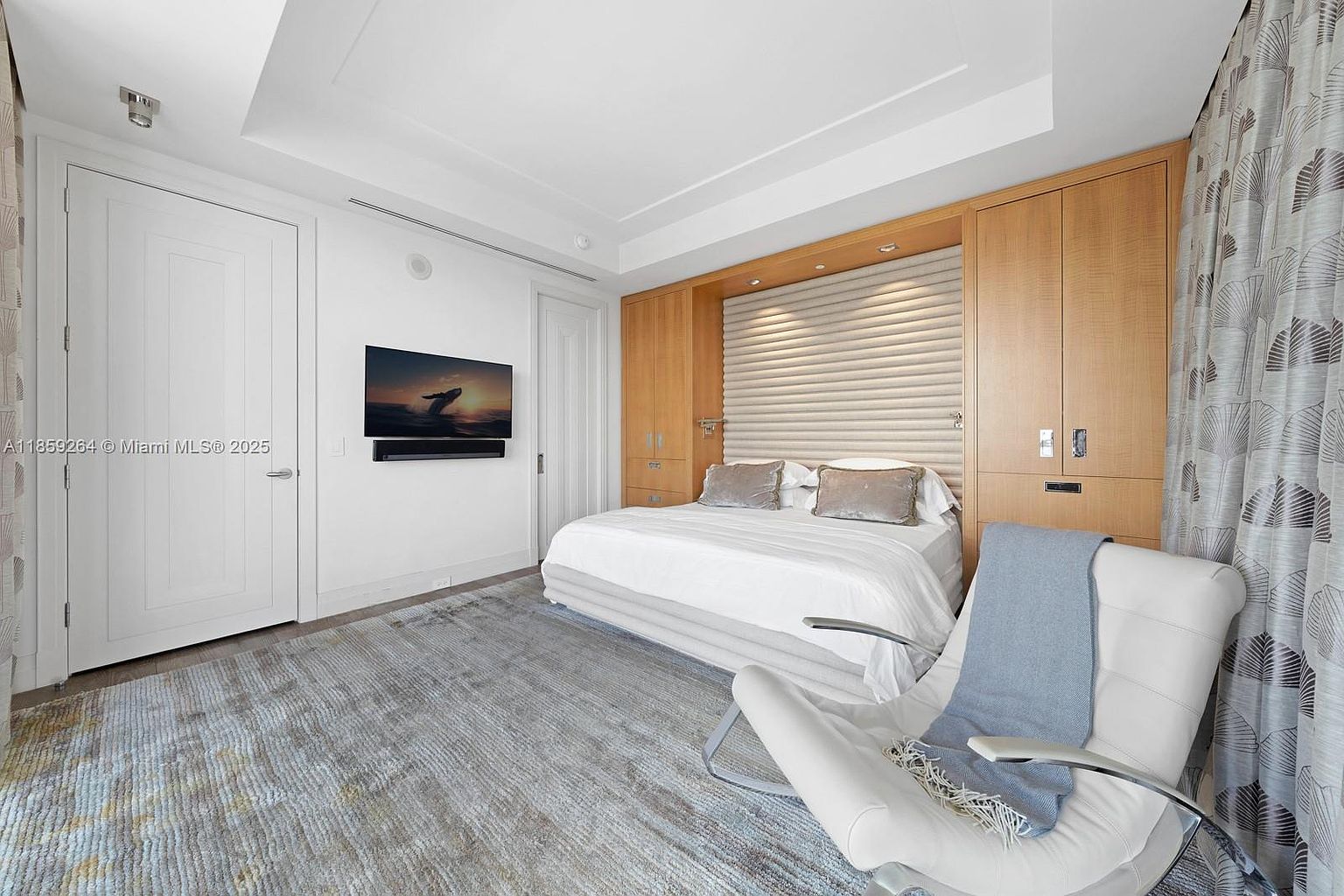This is a well-lit primary bedroom featuring a built-in headboard with integrated lighting and storage. The room includes a large bed with white linens, a modern white chair with a throw blanket, and a flat-screen TV mounted on the wall. The flooring is a patterned area rug, and the overall aesthetic is clean, modern, and luxurious.