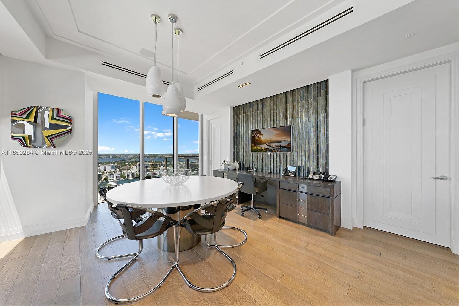 This interior shot showcases a modern dining area with a round white table surrounded by stylish chairs with chrome frames. A nearby desk area features a mounted television and sleek cabinetry, creating a functional and aesthetically pleasing space. Large windows offer a stunning view, enhancing the room's appeal.