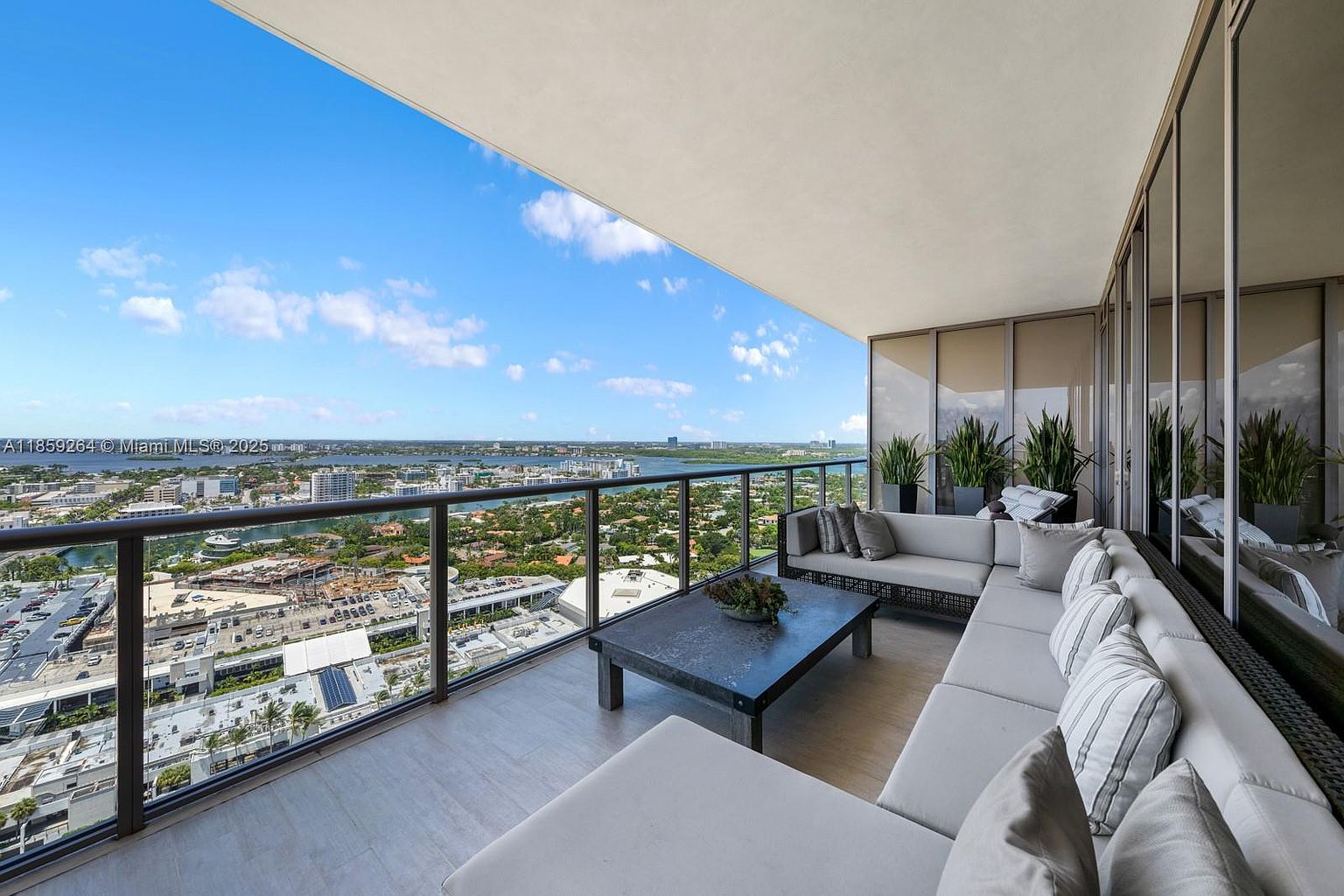 This image showcases a luxurious balcony with a stunning panoramic view of the city and waterfront. The balcony features a comfortable sectional sofa with plush cushions, a modern coffee table, and potted plants, creating an inviting outdoor living space. The glass railing provides an unobstructed view, enhancing the appeal of this high-end property.