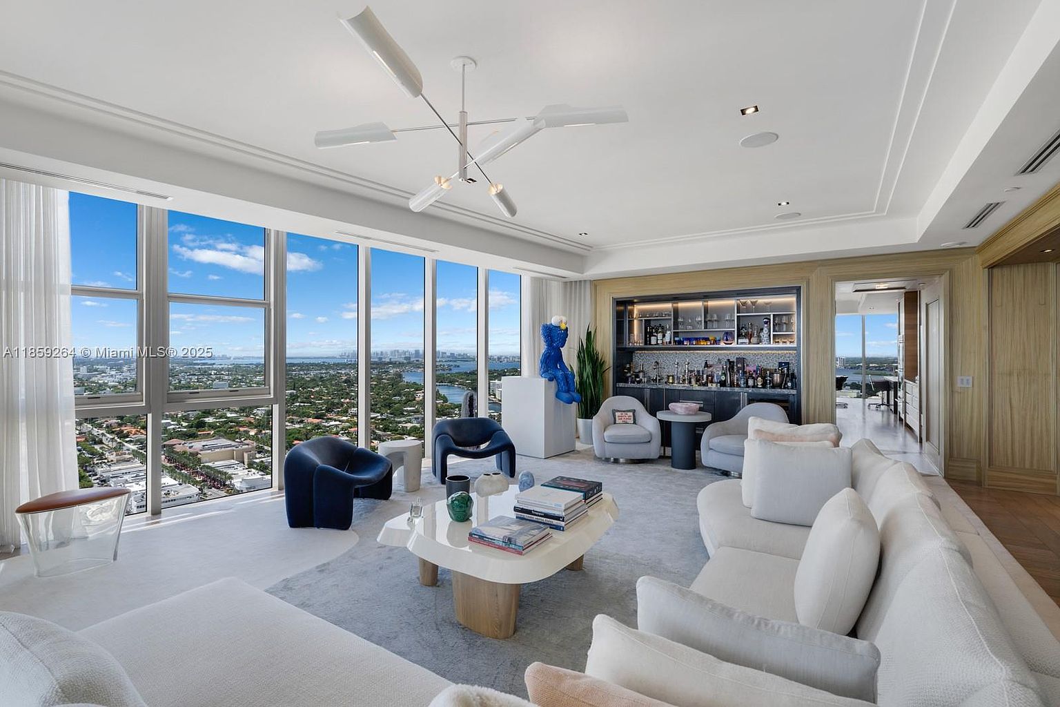 This is a luxurious living room featuring floor-to-ceiling windows with a stunning city view. The room is furnished with a large white sectional sofa, modern armchairs, and a unique coffee table. A built-in bar area adds a touch of sophistication, and the overall design is contemporary and elegant.