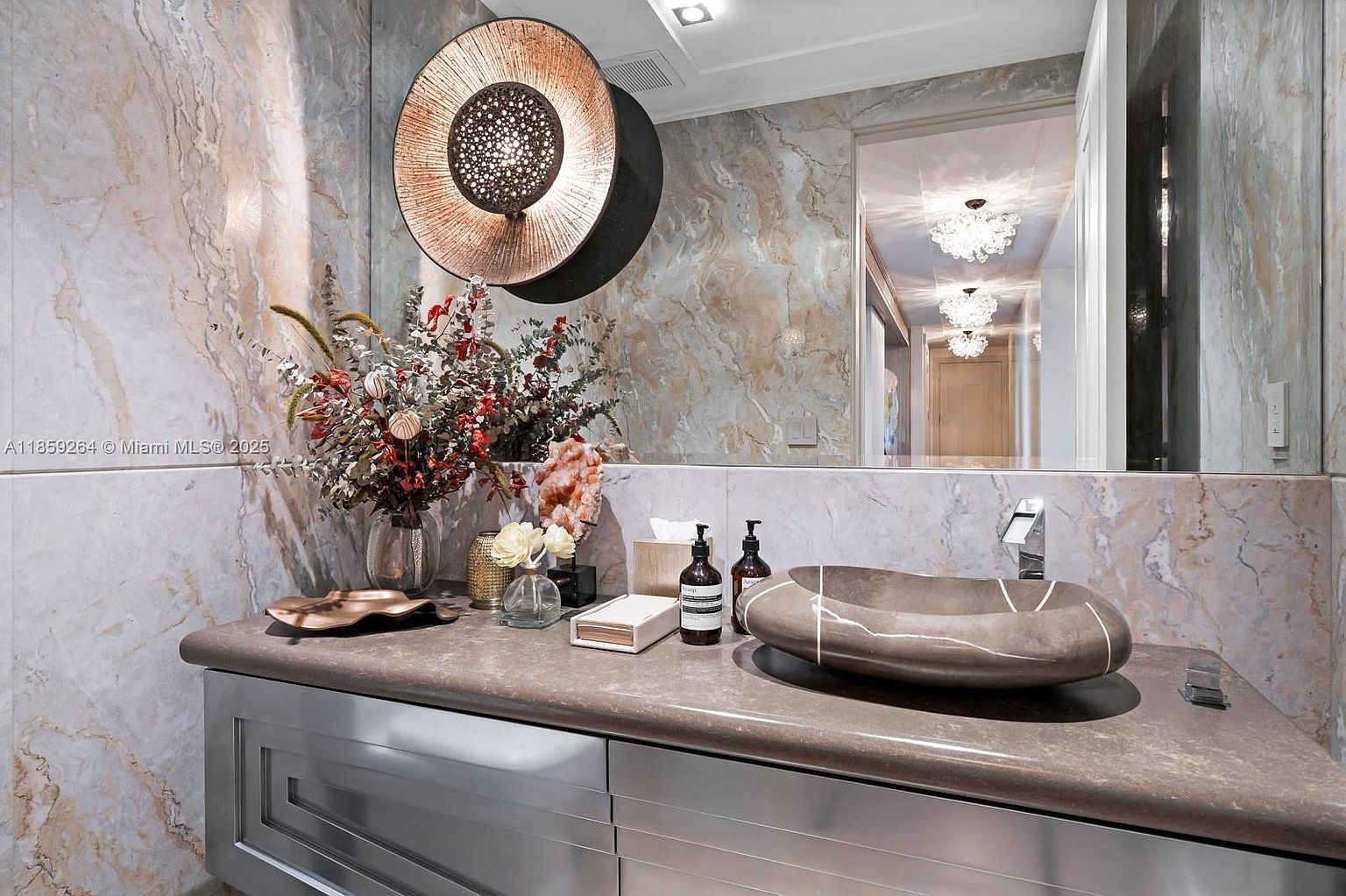 This is a stylish bathroom featuring marble walls and a unique stone sink. A round, modern light fixture hangs above the vanity, which is adorned with decorative items and toiletries. The overall impression is luxurious and contemporary, perfect for a high-end real estate listing.