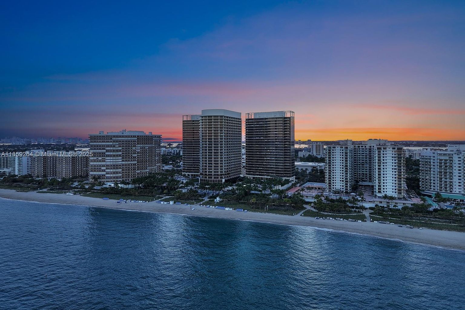 This aerial view showcases a row of high-rise beachfront condominiums at sunset. The buildings are modern and appear luxurious, with direct access to a sandy beach and the ocean. The sky is painted with vibrant colors, enhancing the appeal of this coastal property.